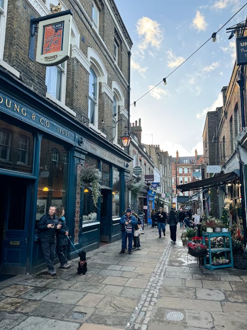 A narrow urban street scene with several pedestrians walking along the paved pavement. On the left, there is a dark blue shopfront with large glass windows displaying inside lighting, situated beneath a white building with arched windows and decorative brickwork. Above the shop, a hanging sign with a red and white design is visible, along with exterior wall-mounted lanterns illuminating the area. On the right, there are small storefronts and outside display areas with potted plants and flowers arranged on racks and tables. Overhead, string lights are strung across the street, adding a festive atmosphere. The sky is partly cloudy with some blue visible. The overall setting appears to be in a lively, commercial area within Hampstead, where alternative waste handling or private rubbish collection might be considered due to the busy street environment and limited space for collections directly from the pavement. Waste Disposal Hampstead often manages collection in similar narrow streets, balancing local authority regulations with private waste removal services.