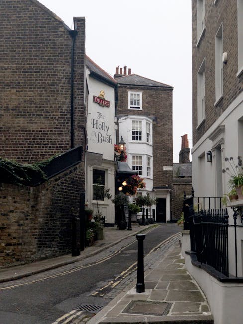 A narrow urban street scene featuring a small, gently curved paved alleyway bordered by brick and stucco buildings. On the left side, a brick wall with a slight overhang and ivy partially covering it runs alongside the alley, leading to a building with the signage for 'The Holly Bush' pub, which has white exterior walls, a bay window, and hanging flower baskets near its entrance. To the right, a white building with a black metal railing and potted plants decorates the front. The street is paved with tarmac and stone, with yellow double lines marking the edge, and a small black bollard stands in the foreground, possibly indicating restricted access or an obstruction for pedestrian or vehicle traffic. Overhead, the sky is overcast, creating a soft, diffused light across the scene, which highlights the textures of the brickwork, window frames, and street surface. Typically, this type of setting relates to independent pubs or small local venues that may rely on private waste disposal services like Waste Disposal Hampstead for rubbish collection, especially considering the narrow access and constrained street layout, which can delay or complicate regular rubbish van visits from larger municipal collection routes.