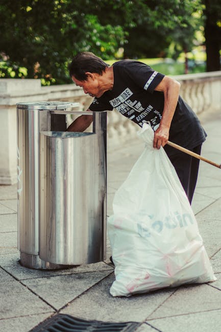 A middle-aged man with dark hair and light skin is seen disposing of waste into a public metal litter bin situated on a paved sidewalk. He is wearing a black T-shirt with white text printed on it, along with dark trousers. The man is bent slightly forward as he places a white plastic bag filled with rubbish into a large, cylindrical, stainless steel bin that has an open top. In his left hand, he holds onto the bag, while his right hand is reaching towards the bin. Behind him, there is lush greenery with trees and bushes, and a low stone or concrete balustrade runs along the background edge. The scene appears to be outdoors during daytime, with natural light illuminating the area, suggesting an environment where public waste disposal is available. The image subtly supports the context of rubbish removal and waste management services, such as those offered by Waste Disposal Hampstead, highlighting individual responsibility for proper waste disposal in public spaces.