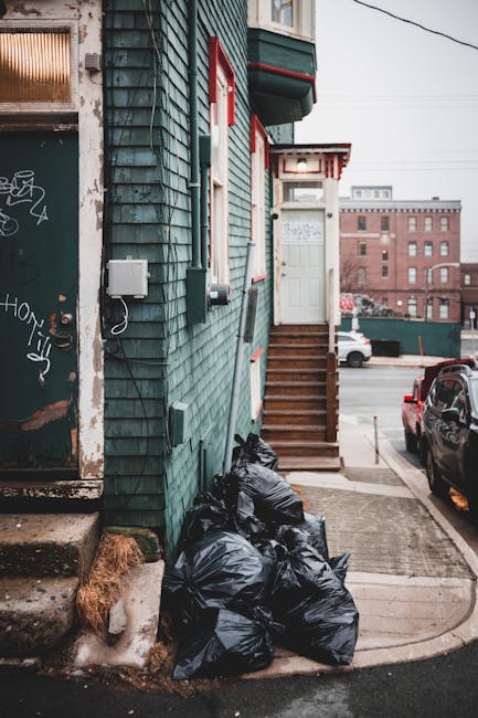 A small urban property with dark green wooden cladding on its exterior wall, positioned on a street corner. A set of wooden stairs with a handrail leads up to a white door with red accents and a small canopy overhead. To the left of the stairs, several tightly packed black rubbish bags are stacked on the concrete sidewalk, partially covering a patch of dried grass near the building's foundation. Nearby, part of an aged metal door with graffiti marks and a weathered surface is visible, along with an electrical box mounted on the wall. In the background, parked cars and a large red brick building across the street are seen under an overcast sky, suggesting a typical residential or mixed-use urban environment. The scene appears to illustrate an example of private waste disposal arrangements, possibly managed by Waste Disposal Hampstead, highlighting the importance of proper rubbish collection for property maintenance and cleanliness.