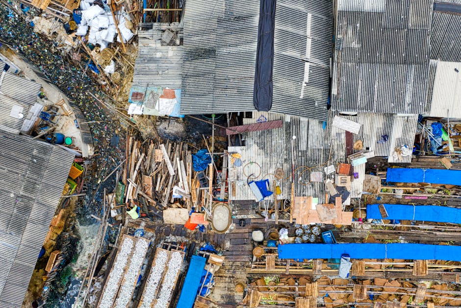 An aerial view of a densely cluttered outdoor area with makeshift structures and sheds constructed from corrugated metal sheets, some rusted and weathered, with visible gaps and irregular overlaps. Piles of discarded wooden planks, broken pallets, and assorted debris are scattered across the space, with some stacked irregularly and others haphazardly placed. The ground features a mixture of concrete, soil, and scattered rubbish, including plastic bottles, metal cans, and other waste materials, indicating a site used for informal waste storage or disposal. Several blue tarp coverings are draped over portions of the debris, possibly to protect or contain specific areas. A narrow pathway runs through the mess, leading to the structures, and various small objects such as buckets, containers, and tools are visible among the clutter. The scene is illuminated by natural daylight, emphasizing the disorderly arrangement and the variety of textures and colours, aligning with private waste collection or onsite clearance services like those offered by Waste Disposal Hampstead in managing local rubbish and debris removal.