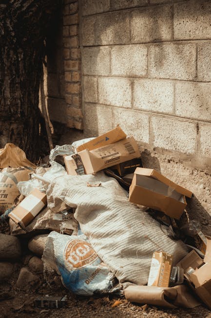 A small urban property with dark green wooden cladding on its exterior wall, positioned on a street corner. A set of wooden stairs with a handrail leads up to a white door with red accents and a small canopy overhead. To the left of the stairs, several tightly packed black rubbish bags are stacked on the concrete sidewalk, partially covering a patch of dried grass near the building's foundation. Nearby, part of an aged metal door with graffiti marks and a weathered surface is visible, along with an electrical box mounted on the wall. In the background, parked cars and a large red brick building across the street are seen under an overcast sky, suggesting a typical residential or mixed-use urban environment. The scene appears to illustrate an example of private waste disposal arrangements, possibly managed by Waste Disposal Hampstead, highlighting the importance of proper rubbish collection for property maintenance and cleanliness.