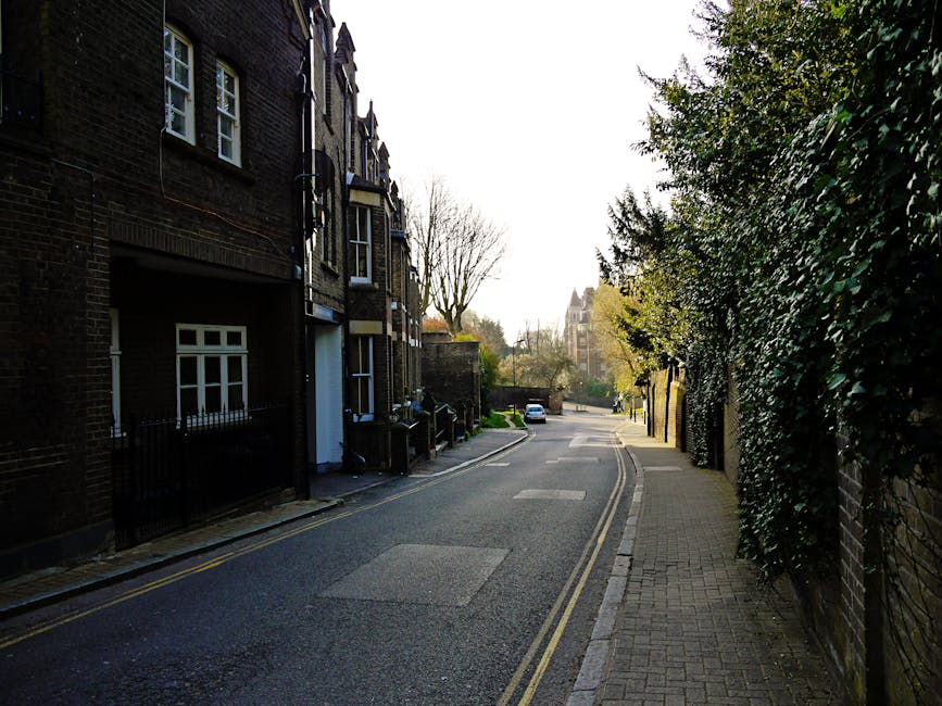 A narrow residential street with a paved asphalt road and a brick-paved sidewalk, lined with dark brick terraced houses on the left side, each featuring white-framed windows and small gates or fences at their entrances. On the right side, there is a tall, dense hedge with dark green, glossy leaves growing along a brick wall, partially obscuring it. The street curves gently to the left in the distance, where a white car is parked, and a church or similar historic building with a pointed tower is visible further along. The scene is illuminated by soft, natural daylight, with some shadows cast by nearby trees, which are mostly leafless, indicating late autumn or early winter. The environment appears clean and orderly, with no visible litter or debris, and the setting suggests a quiet, well-maintained neighborhood suitable for private waste disposal or alternative rubbish collection, such as independent rubbish removal services like Waste Disposal Hampstead, operating in the vicinity.