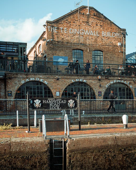 The image depicts a historic brick building named 'The Tedingwall Building', featuring a large painted sign on the upper facade. Situated in an urban environment, the building has at least two levels, with the upper level accessible via exterior metal stairs. Black metal fencing surrounds the ground-floor area, which is marked with signs for 'Camden Lock' and 'Hampstead Road Lock' and features a sign indicating the Regent's Canal. Several pedestrians are visible walking along the sidewalk next to the building, some engaged in conversation, and others passing by. The background shows a partly cloudy sky with blue tones, contributing to a clear daytime scene. The scene captures the area as a bustling spot that might involve private waste handling or local rubbish collection, consistent with services like those provided by Waste Disposal Hampstead. The combination of historic industrial architecture and modern activity suggests ongoing use of the site for leisure or commerce, with the fencing and signage emphasizing its designated public and possibly maintenance-related functions.