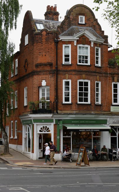 The image depicts a three-story red brick building on a city street, with a bakery, café, and pâtisserie on the ground floor. The building features large white-framed windows, some with small black iron railings, and a decorative brickwork façade with a curved parapet at the top. The bakery storefront has a green awning with white lettering, and a wooden sandwich board sign is placed on the sidewalk outside. In the foreground, a few pedestrians are walking past, with some seated outside the café area. The scene is illuminated by natural daylight, and surrounding trees and neighboring buildings are partially visible, contributing to an urban streetscape ambiance. This setting illustrates a typical downtown area where waste disposal services, such as those offered by Waste Disposal Hampstead, might assist with rubbish collection or on-site clearance, especially for businesses and cafés handling daily waste. The image emphasizes an active commercial environment with potential needs for efficient rubbish removal in busy town areas.