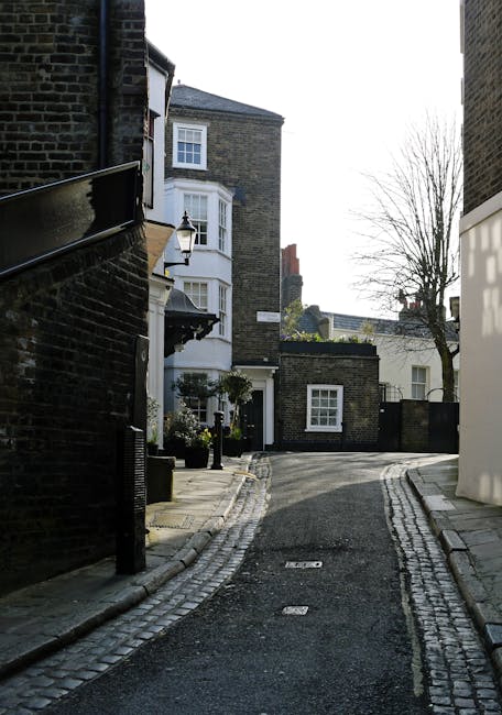 The image depicts a historic brick building named 'The Tedingwall Building', featuring a large painted sign on the upper facade. Situated in an urban environment, the building has at least two levels, with the upper level accessible via exterior metal stairs. Black metal fencing surrounds the ground-floor area, which is marked with signs for 'Camden Lock' and 'Hampstead Road Lock' and features a sign indicating the Regent's Canal. Several pedestrians are visible walking along the sidewalk next to the building, some engaged in conversation, and others passing by. The background shows a partly cloudy sky with blue tones, contributing to a clear daytime scene. The scene captures the area as a bustling spot that might involve private waste handling or local rubbish collection, consistent with services like those provided by Waste Disposal Hampstead. The combination of historic industrial architecture and modern activity suggests ongoing use of the site for leisure or commerce, with the fencing and signage emphasizing its designated public and possibly maintenance-related functions.