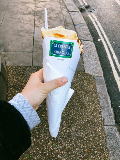 A close-up of a person's hand holding a wrapped takeaway crepe from 'La Creperie de Hampstead,' with a plastic fork inserted into the folded pastry. The crepe has a golden-brown surface and is partially visible above the white paper wrapping featuring the cafe's green and blue logo and text indicating its location in Hampstead. The background shows a paved pedestrian walkway with a textured surface of small stones and concrete slabs, and a street edge with asphalt, white double yellow lines, and a drain cover. The scene appears to be outdoors in a street setting, with natural daylight illuminating the crepe and surrounding environment, relevant to outdoor food vendors that may generate waste requiring collection services from companies like Waste Disposal Hampstead, which handles rubbish removal in the area.