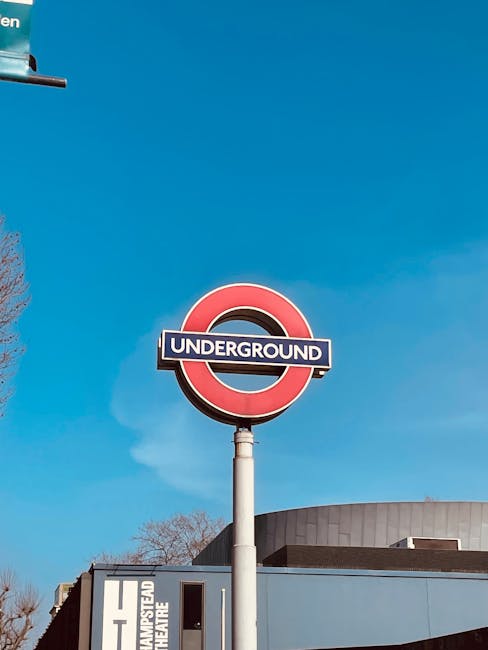A circular London Underground sign featuring a red outer ring and a blue bar with the word 'UNDERGROUND' in white, mounted on a white pole against a bright blue sky with some faint clouds. In the background, part of a modern building with a curved, grey roof and a blue facade is visible, with a sign indicating Hampstead Theatre. The scene is outdoors near the Hampstead area, with leafless trees partially visible on the left side. This image emphasizes urban infrastructure and public transport signage, relevant to alternative waste handling discussions by highlighting city environment features. The sign's placement and clarity make it a recognizable icon of London's underground network, often associated with city-wide mobility and infrastructure support, indirectly connected to services like rubbish collection and disposal logistics managed by waste disposal specialists such as Waste Disposal Hampstead.