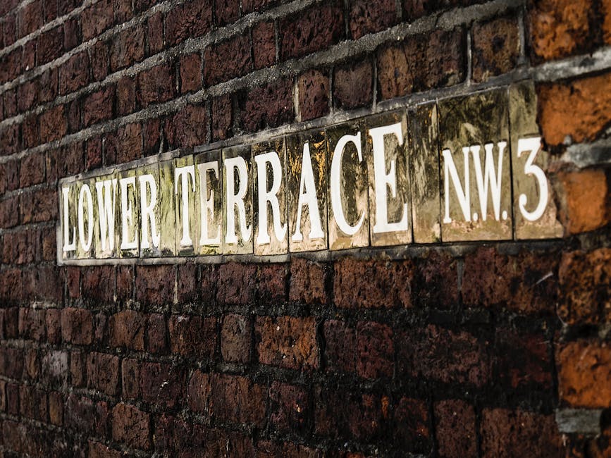 A close-up view of a rectangular street sign mounted on a weathered red brick wall, displaying the words 'LOWER TERRACE NW3' in large, white, capital letters. The sign appears to be made of ceramic tiles with a glossy surface, some of which show signs of moss and dirt accumulation, particularly around the edges. The brick wall consists of dark reddish-brown bricks with a rough texture, arranged in a traditional running bond pattern, and mortar joints that are slightly recessed. The sign is positioned horizontally along the wall, with the bricks serving as a textured background. The overall scene is illuminated with natural light, highlighting the aged and rustic appearance of both the wall and the sign, which contributes to an atmospheric urban setting typical of older residential or public areas. This image visually supports themes related to urban environments, historical signage, or heritage features, aligning subtly with private property markers or street identification for services like rubbish removal or property maintenance in the area.