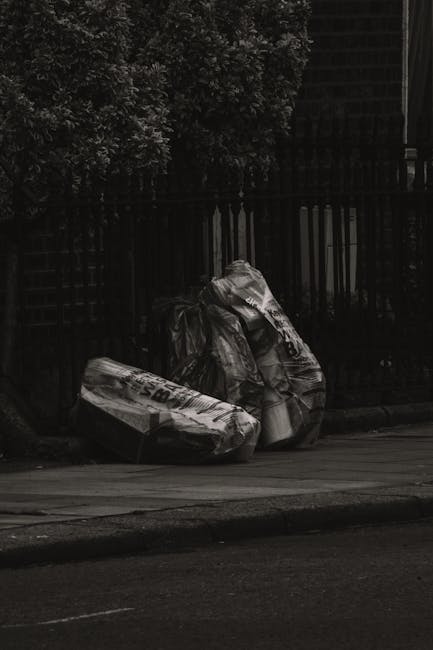 Two large black plastic rubbish bags, slightly crumpled and protruding at the top, are placed on a paved sidewalk beside a dark metal fence with vertical bars. The bags appear to contain waste, possibly household refuse, and are partially obscuring what looks like an area of dense foliage and trees behind the fence. The scene is set outdoors during daytime, with soft natural lighting casting gentle shadows on the pavement, and the overall atmosphere suggests a temporary rubbish collection point. The setting aligns with private waste disposal practices, as it shows bags prepared for collection rather than curbside bin services, reflecting an approach to independent rubbish removal commonly managed by services like Waste Disposal Hampstead. The environment appears clean aside from the waste bags, indicating a routine rubbish disposal area near a residential or urban property, highlighting the importance of organized waste removal for maintaining cleanliness and adhering to local or private disposal protocols.